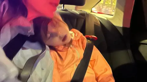 Contribuido a la niña durmiendo en la parte trasera del auto. Su cabeza descansa sobre los hombros de la mujer. Ella usa un abrigo naranja y tiene un cinturón de seguridad.