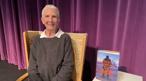 BBC Ann Cleeves, pelo corto y blanco y sonrisas en sillas de madera tejida. Lleva una camisa gris oscuro sobre una camisa de cuello blanco. Detrás de ella hay un escenario con un velo de color rosa intenso. Su nuevo libro a su lado es una piedra sobre una pequeña mesa blanca. 