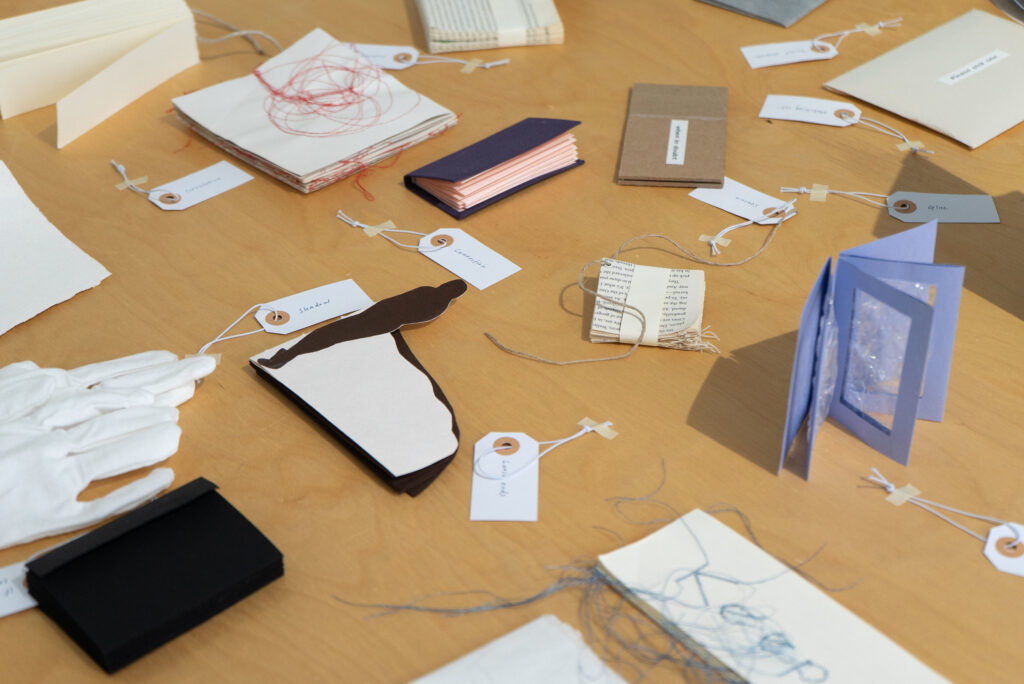 Una mesa de madera con libros hechos a mano, manualidades en papel, guantes, hilos enredados y pequeñas etiquetas con etiquetas escritas a mano sugiere un proyecto artístico o de archivo.