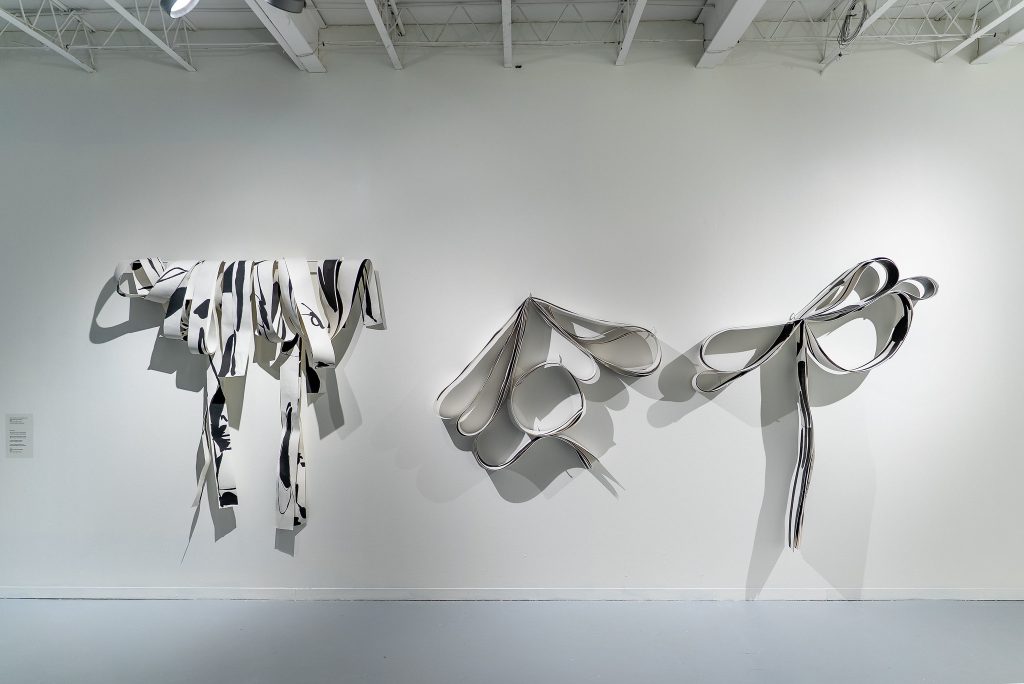 Tres esculturas abstractas en blanco y negro están colocadas contra una pared blanca de la galería hecha de cinta doblada y rizada, suavemente sombreada. Las formas son similares a cintas drenadas o enrolladas.