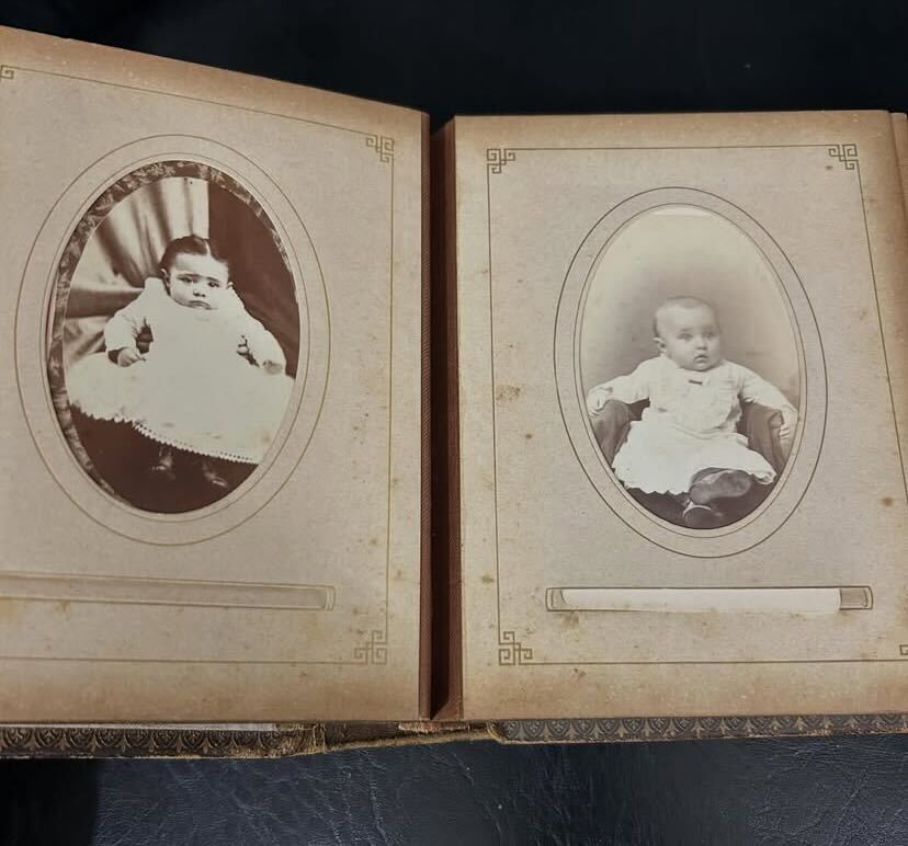 Un álbum de fotos abierto presenta dos retratos de un bebé con una bata blanca enmarcados por un fondo ovalado en una página envejecida.